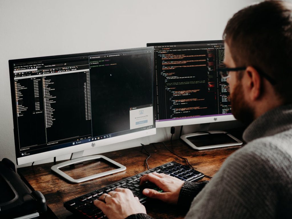 pexels-photo-16129724 Software developer typing code on dual monitors at a wooden desk.